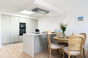 Eine moderne Küche mit integrierten Geräten, ein Essbereich mit rundem Tisch und Korbstühlen sowie Meerblick durch das Fenster.