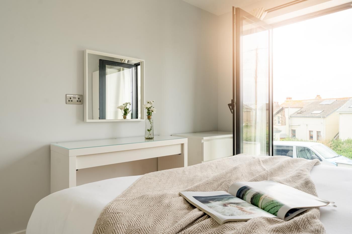 A bedroom with a bed, a bedside table, a mirror, and a window with a view of a car and buildings outside.