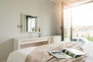 A bedroom with a bed, a bedside table, a mirror, and a window with a view of a car and buildings outside. - 2 Bed in Bude (oc-b588787) (Bude)