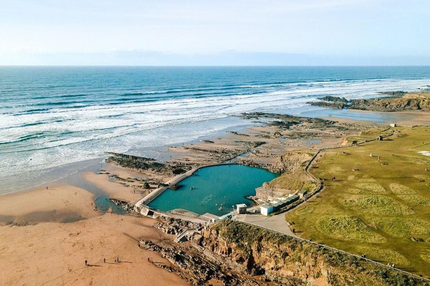 Luftaufnahme einer Küstenlandschaft mit einer weiten Wasserfläche, felsigem Gelände und einem eingezäunten Bereich mit Gebäuden.