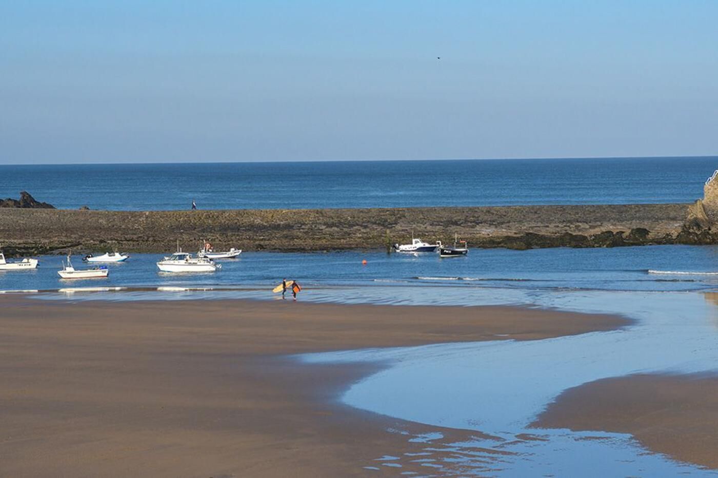 A beach with boats, a rocky shoreline, and a person with a surfboard.