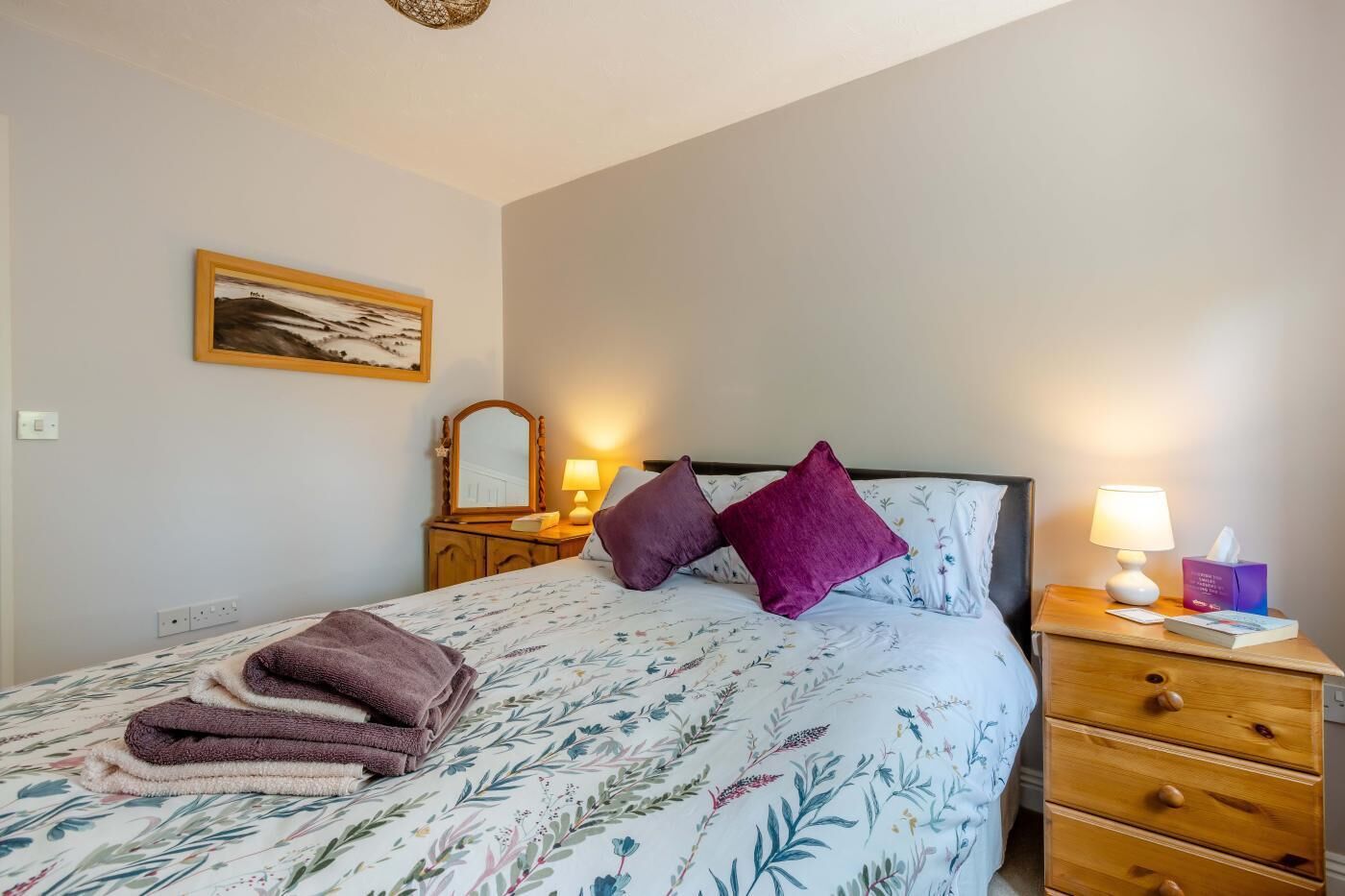 Ein ordentlich bezogenes Bett mit Blumendecke, zwei Kissen, einer Nachttischlampe und einem kleinen Nachttisch aus Holz mit Lampe und Taschentuchbox.