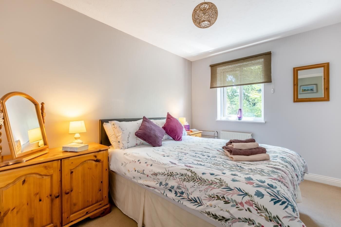 A bedroom with a bed, a wooden dresser, a mirror, a window with a view, and a ceiling light fixture.