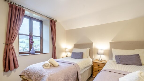 A bedroom with two beds, a window with curtains, a nightstand with lamps, and a view of the outside. - 5 Bed in Grizebeck (oc-b34029) (Kirkby Ireleth)