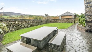 A stone patio with a rectangular table and benches, surrounded by a stone wall and a grassy area. - 5 Bed in Grizebeck (oc-b34029) (Kirkby Ireleth)