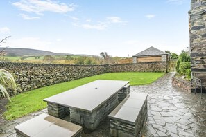 A stone patio with a rectangular table and benches, surrounded by a stone wall and a grassy area. - 5 Bed in Grizebeck (oc-b34029) (Kirkby Ireleth)