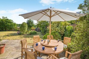 A round wooden table with six chairs and a large umbrella, surrounded by lush greenery and a stone pathway. - 3 Bed in Briningham (oc-s44878) (Briningham)