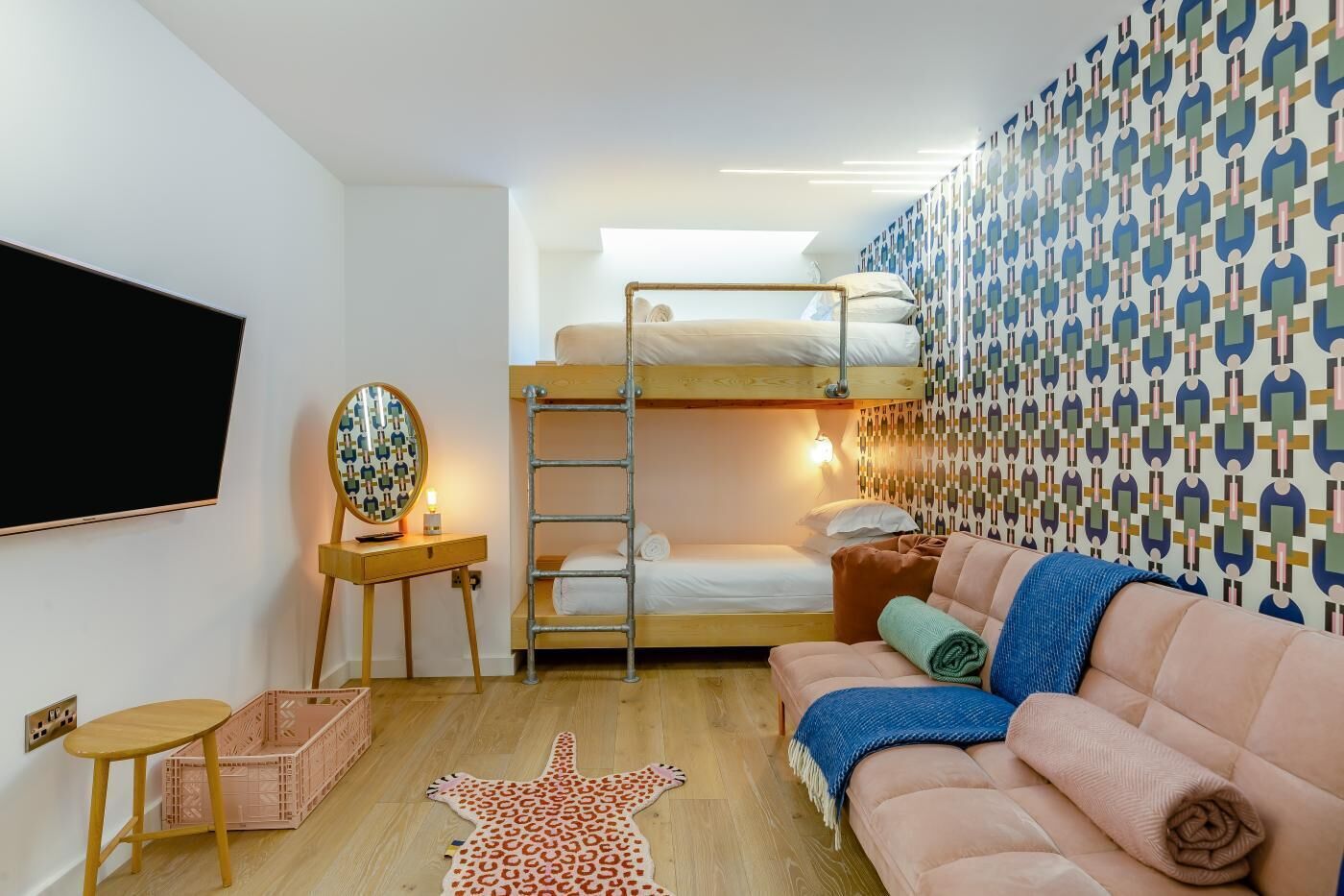 A bunk bed room with a television, a desk with a mirror, a patterned wall, and a sofa.