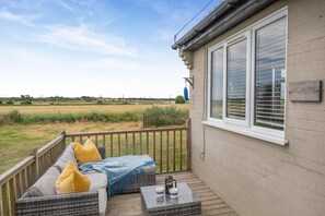 A covered outdoor seating area with wicker furniture and a table, overlooking a field. - 1 Bed in Eccles-on-Sea (oc-w45218) (Lessingham)
