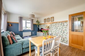 A cozy room with a sofa, a dining table with chairs, a TV, and a window with curtains. - 1 Bed in Eccles-on-Sea (oc-w45218) (Lessingham)