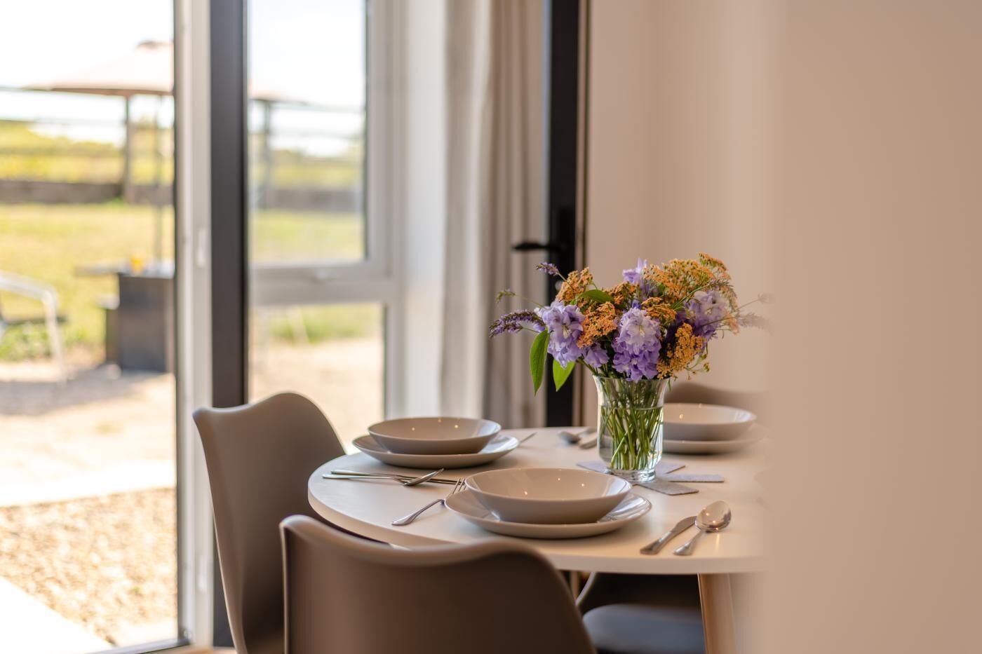 Et spisebord dækket med tallerkener, bestik og en vase med blomster, med udsigt til det fri via store vinduer.