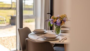 Mesa de jantar posta com pratos, talheres e um vaso de flores, com vista para o exterior através de grandes janelas.