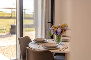 A dining table set with plates, cutlery, and a vase of flowers, with a view of the outdoors through large windows. - 1 Bed in Birchington (oc-s45228) (Birchington-on-Sea)