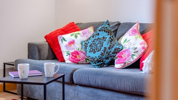 Um sofá cinzento com almofadas florais e estampadas, uma mesinha com uma caneca branca e um caderno rosa, e um tapete vermelho e cinzento.