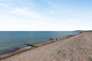 Ein Kiesstrand mit ruhiger See, einer Holzsteganlage und einer entfernten Küstenlinie mit Gebäuden.