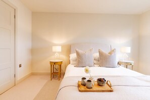 A hotel room with a large bed, two bedside tables with lamps, a tray with cups and a flower, and a door on the left. - 3 Bed in Watergate Bay (oc-b642096) (Tregurrian)