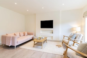 A modern living room with a sofa, a coffee table, a chair, a flat-screen TV mounted on the wall, and a lamp. - 3 Bed in Watergate Bay (oc-b642096) (Tregurrian)