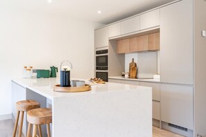 A modern kitchen with a central island, wooden stools, and a variety of appliances. - 3 Bed in Watergate Bay (oc-b642096) (Tregurrian)