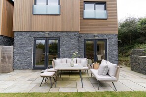 A modern outdoor patio with a dining set, stone wall, and wooden paneling. - 3 Bed in Watergate Bay (oc-b642096) (Tregurrian)