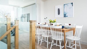 A modern dining area with a wooden table and white chairs, a glass partition, and two framed artworks on the wall. - 4 Bed in Porth (oc-b584252) (Newquay)