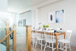 A modern dining area with a wooden table and white chairs, a glass partition, and two framed artworks on the wall.