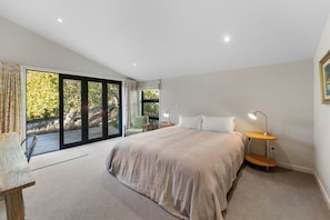 Ein Schlafzimmer mit einem großen Bett, zwei Nachttischlampen, einem Sessel und Blick durch eine Schiebetür in einen Außenbereich.