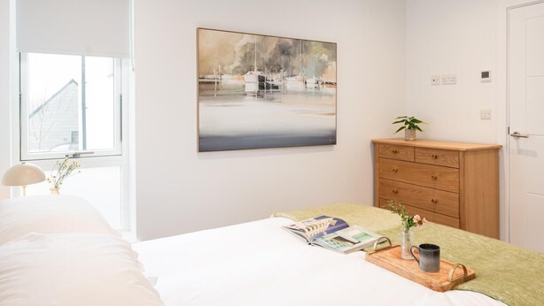 A bedroom with a bed, a bedside table with a book and a vase, a wooden dresser, and a painting on the wall. - 2 Bed in Falmouth (oc-b669230) (Flushing)