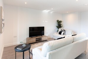 Un salon moderne avec un téléviseur à écran plat, un canapé blanc, une petite table d’appoint et une plante en pot.
