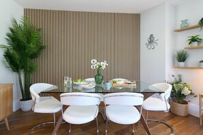 A dining area with a glass table surrounded by white chairs, a vertical wooden panel wall, and shelves with plants. - 2 Bed in Watergate Bay (oc-b584254) (Tregurrian)