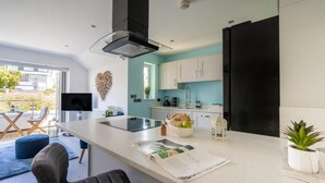 A modern kitchen with a large island, a flat-screen TV, and a dining area with a blue sofa and a chair. - 2 Bed in Porth (oc-b588797) (Newquay)