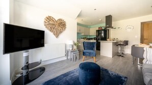 A modern living room with a flat-screen TV, a heart-shaped wall decoration, a blue velvet chair, a dark blue ottoman, and a kitchen area in the background. - 2 Bed in Porth (oc-b588797) (Newquay)