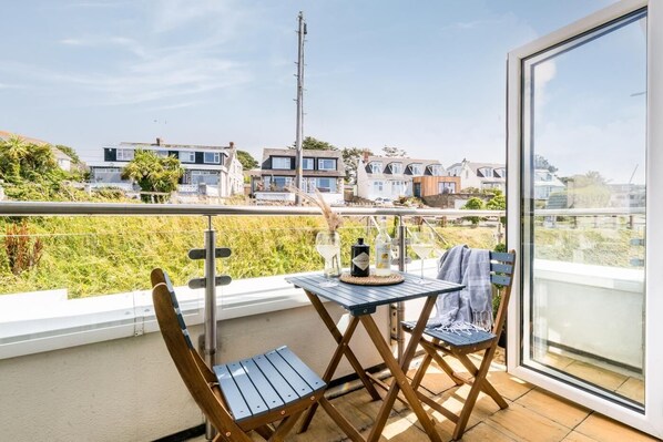 A balcony with a table, chairs, and a bottle of wine, overlooking a residential area with houses and a grassy field. - 2 Bed in Porth (oc-b588797) (Newquay)