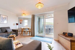 A modern living room with a sofa, a dining table, and a TV mounted on the wall. - 2 Bed in Watergate Bay (oc-b590010) (Watergate Bay)