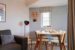 A modern living room with a sofa, a dining table with chairs, a floor lamp, and artwork on the wall. - 2 Bed in Watergate Bay (oc-b590010) (Watergate Bay)