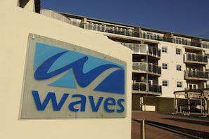 A building with a sign that reads 'Waves' and a multi-story residential structure with balconies in the background. - 2 Bed in Watergate Bay (oc-b590010) (Watergate Bay)