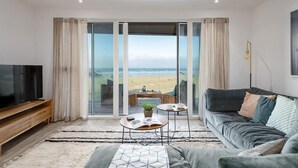 Een moderne woonkamer met een grote televisie, een bank, een salontafel en uitzicht op het strand door glazen deuren.