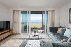 Ein modernes Wohnzimmer mit großem Fernseher, einer Couch, einem Couchtisch und Blick auf den Strand durch Glasüren.