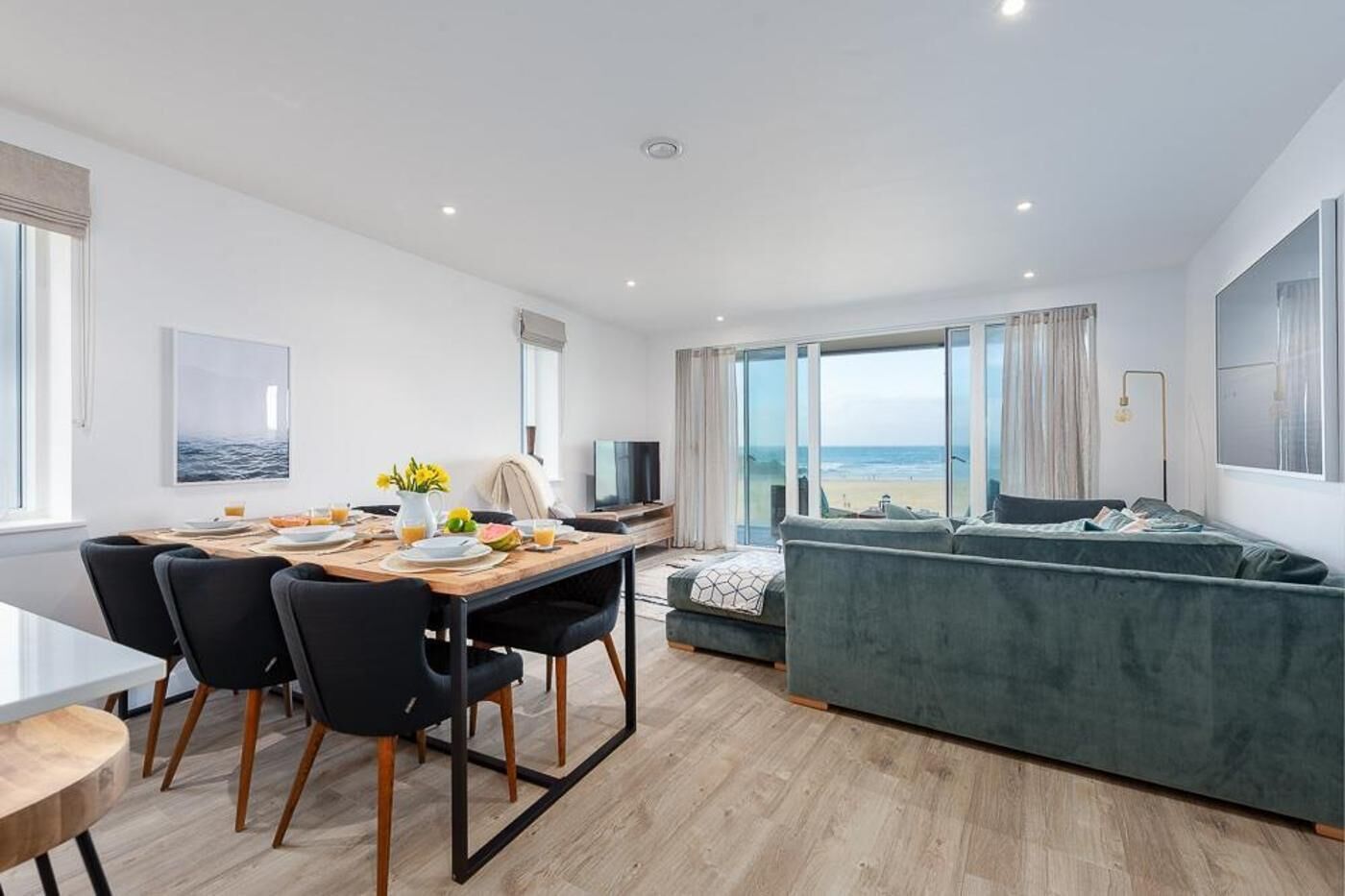 Una sala de estar moderna con un ventanal grande con vista a la playa, una zona de comedor con una mesa y sillas de madera, y un sofá gris.