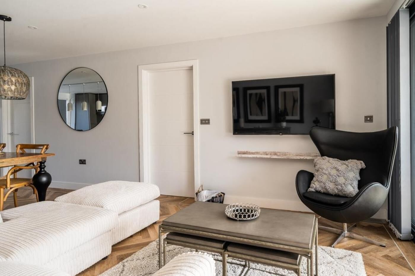 Un salon moderne avec un canapé, une table basse, un fauteuil noir, un miroir rond et un téléviseur à écran plat fixé au mur.