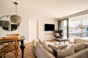 A modern living room with a dining area, a large mirror, a flat-screen TV, and a view of a car through a glass door. - 2 Bed in Bude (oc-b588786) (Bude)