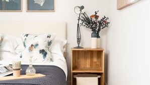 A neatly arranged bedroom with a bed, pillows, a bedside table with a lamp, a vase with flowers, and a shelf with a basket. - 3 Bed in Hayle (oc-b624277) (Hele)