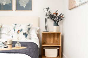 A neatly arranged bedroom with a bed, pillows, a bedside table with a lamp, a vase with flowers, and a shelf with a basket. - 3 Bed in Hayle (oc-b624277) (Hele)