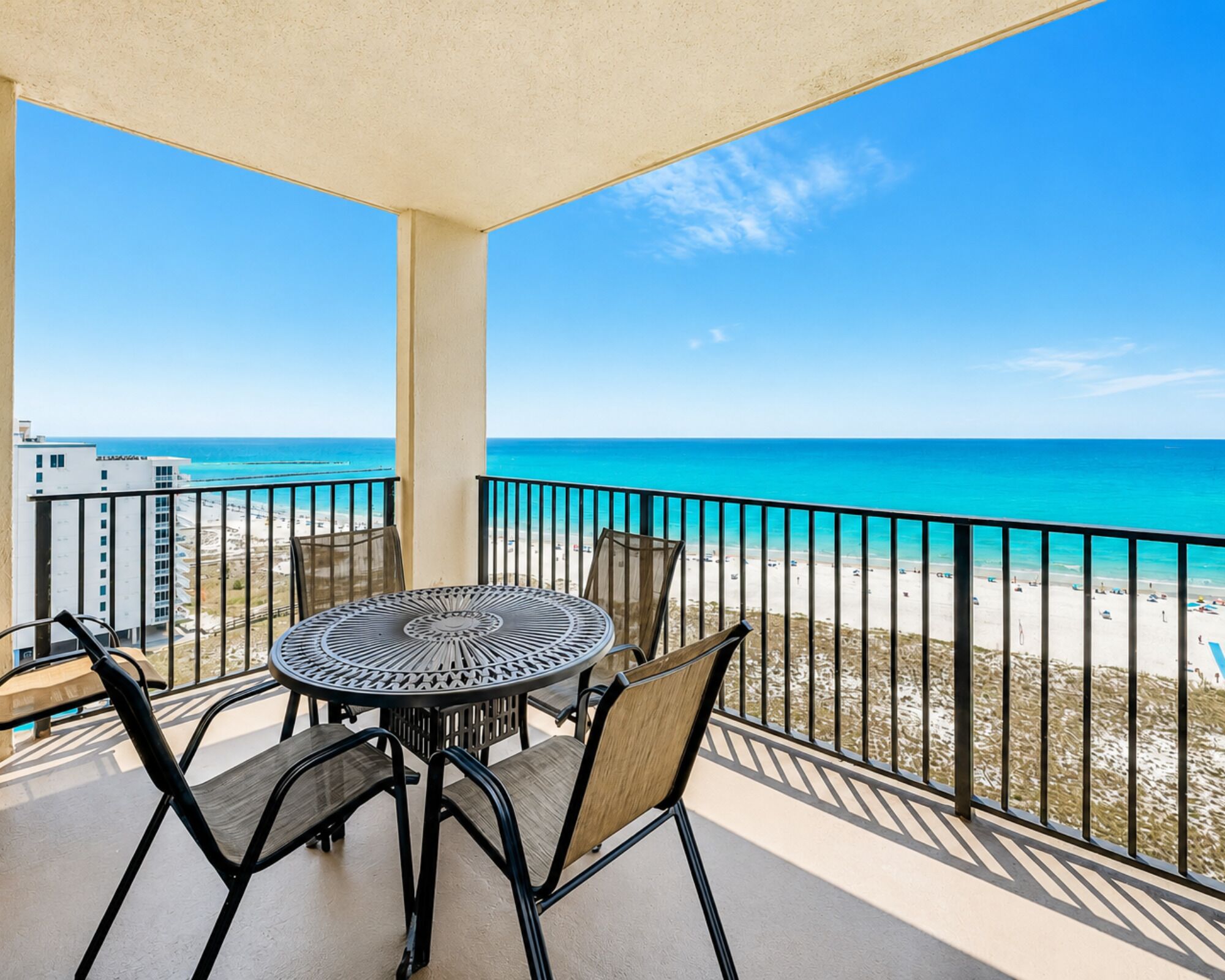 A balcony with a round table and chairs, overlooking a beach with clear blue water and a sandy shore.