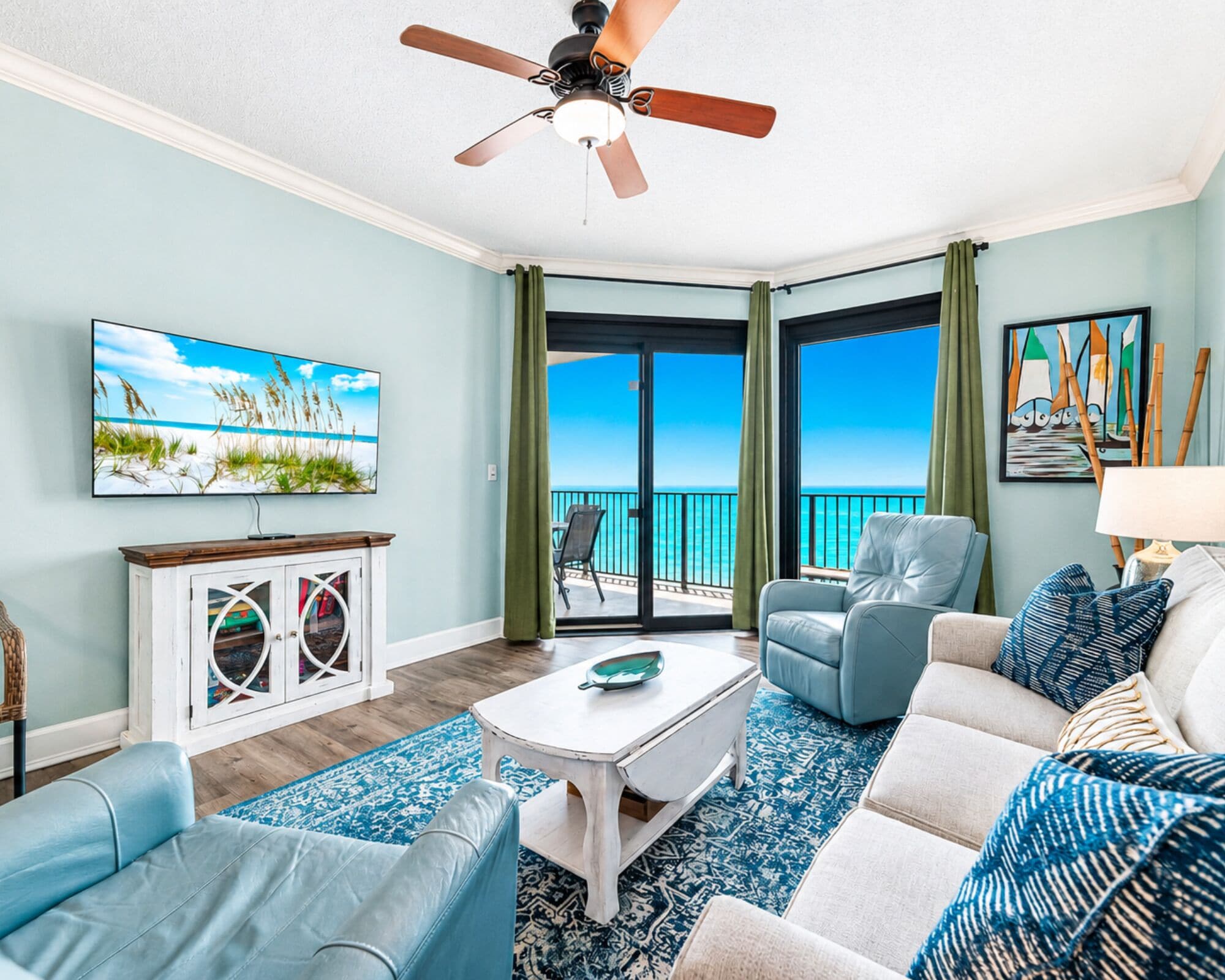 A living room with a ceiling fan, a television, a sofa, a coffee table, and a view of the ocean through a sliding glass door.