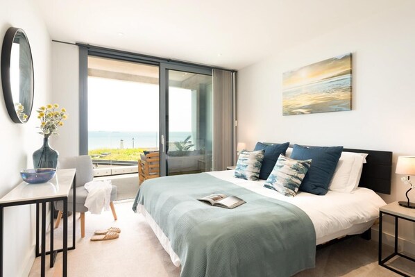 Un dormitorio con cama, escritorio, silla, un florero con flores y vista al mar.