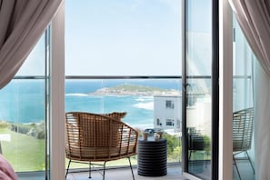 A balcony with a wicker chair and a small round table, overlooking a coastal view with a building in the distance. - 4 Bed in Fistral (oc-b584261) (Newquay)