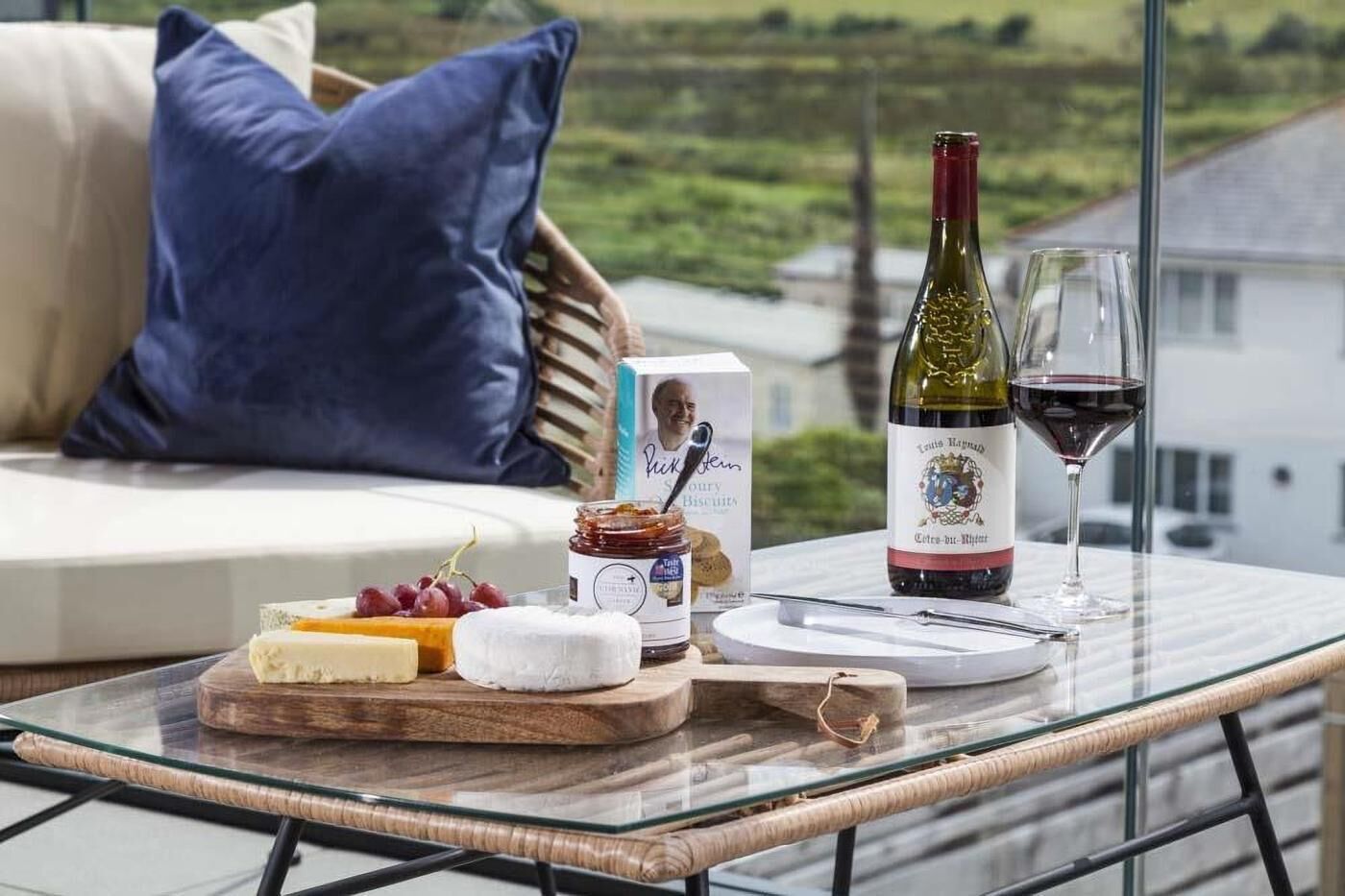 Une table en verre sur laquelle se trouvent une bouteille de vin, un verre de vin, du fromage, des raisins et un pot de confiture, le tout placé sur un balcon d’où l’on peut admirer une maison et de la verdure.