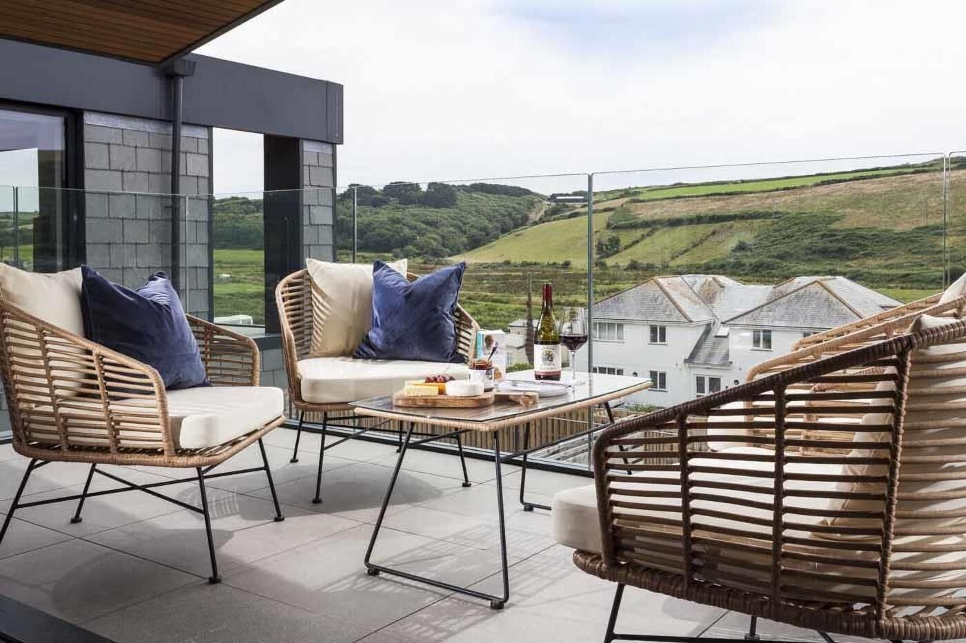 A balcony with wicker furniture, a table with a bottle and glasses, and a view of houses and hills.
