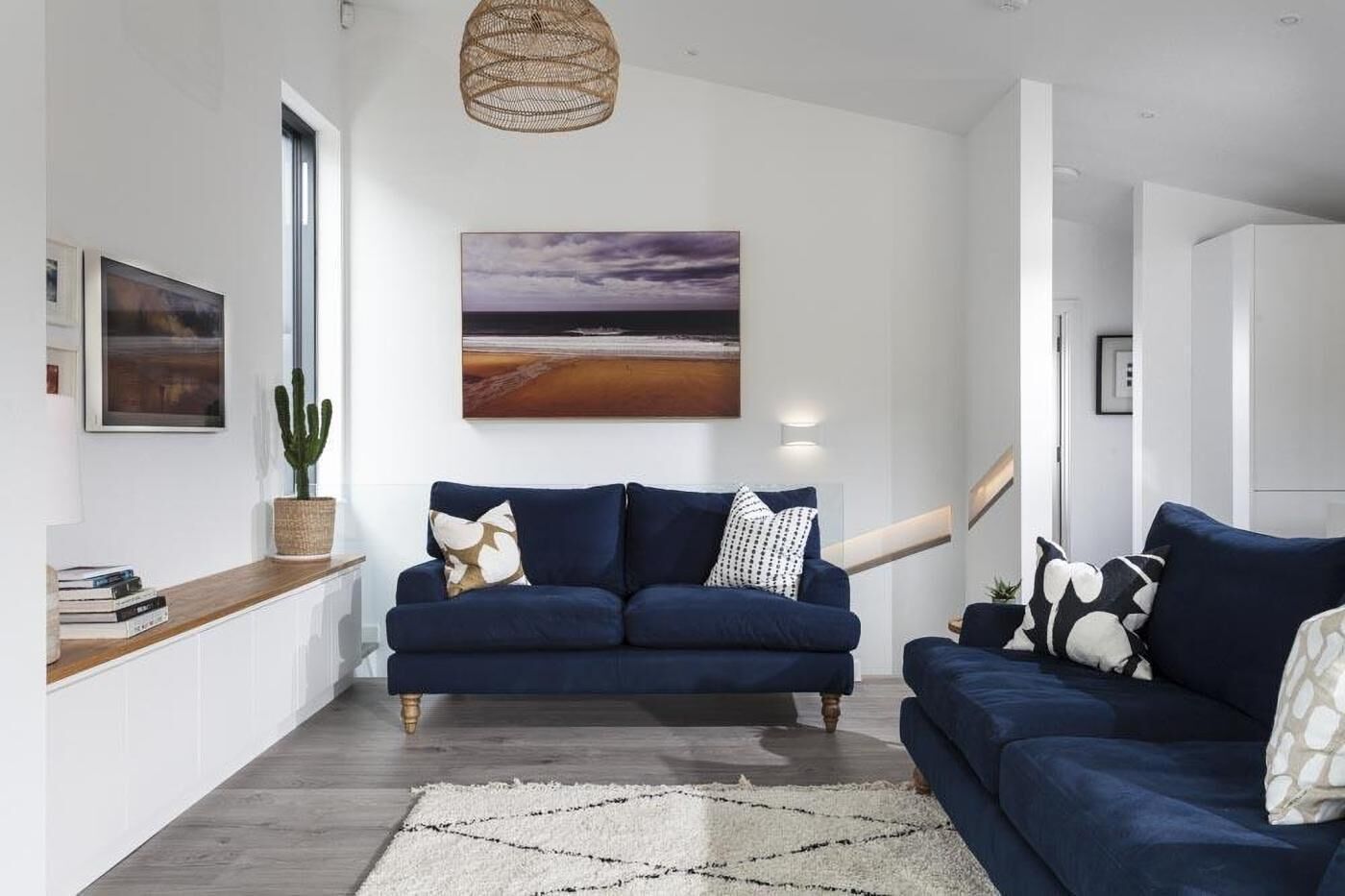 Un salon moderne avec un canapé bleu foncé, un grand tableau représentant un coucher de soleil et un cactus.
