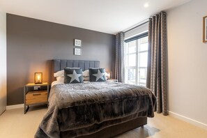 A bedroom with a large bed, a nightstand with a lamp, and a window with curtains. - 4 Bed in Holywell Bay (oc-b659846) (Holywell)